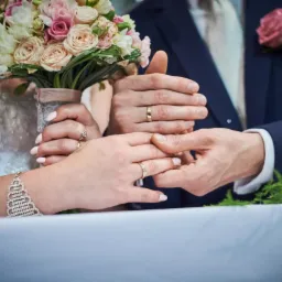 Dłonie nowożeńc&oacute;w z obrączkami na tle bukietu ślubnego i garnituru, subtelne światło podkreśla detale biżuterii.