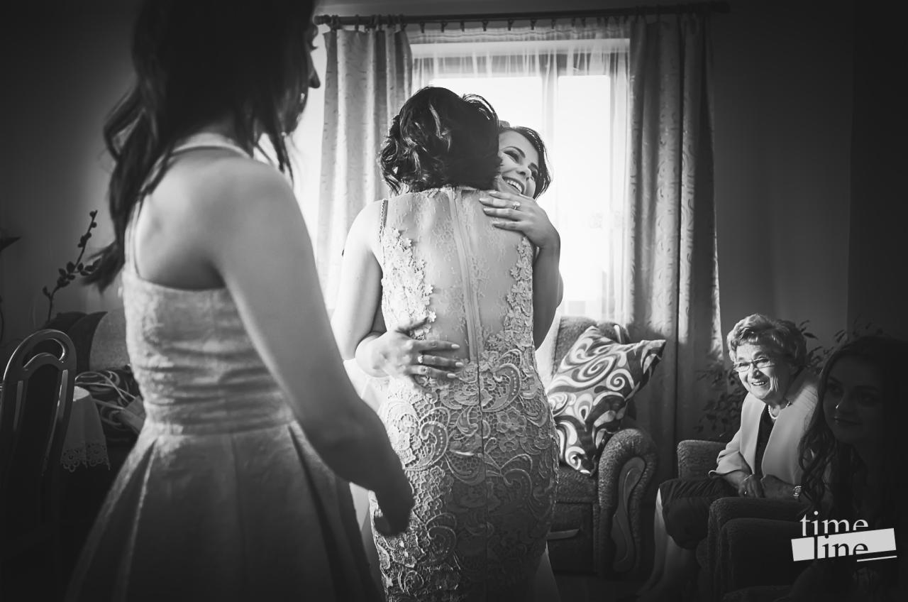 Czarno-białe zdjęcie uśmiechniętej panny młodej w koronkowej sukni obejmującej inną kobietę, z widocznymi gośćmi w tle i oknem z zasłonami.