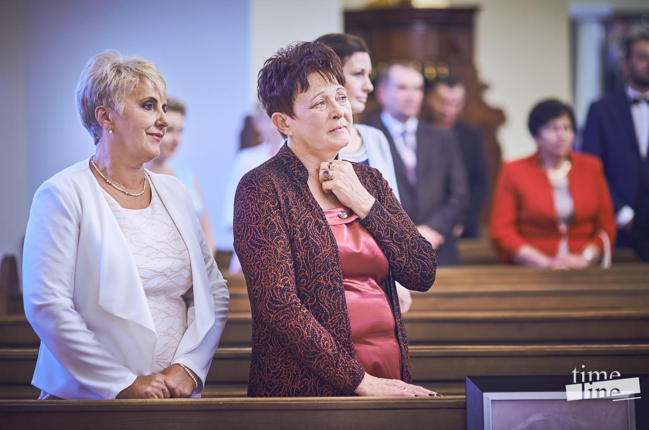 Goście weselni w kościele, kobiety w eleganckich strojach siedzą w ławkach, w tle widoczni inni uczestnicy uroczystości.