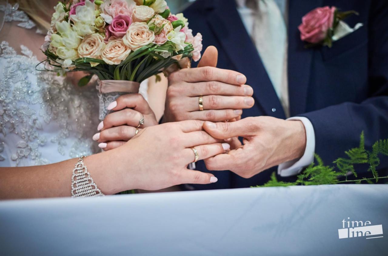 Dłonie nowożeńców z obrączkami na tle bukietu ślubnego i garnituru, subtelne światło podkreśla detale biżuterii.