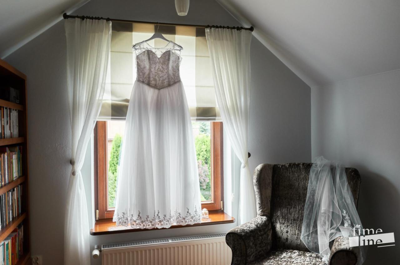 Elegancka suknia ślubna z koronkowym gorsetem i tiulowym dołem, zawieszona na wieszaku w oknie z białymi zasłonami, obok fotel z welonem, w tle widok na ogród.