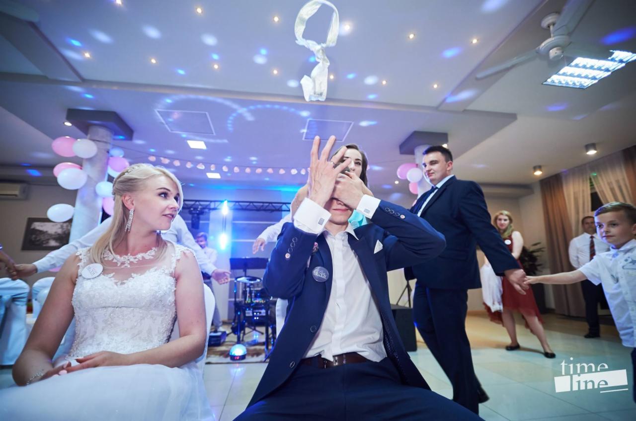 Panna młoda siedzi w sukni ślubnej, podczas gdy pan młody ma zasłonięte oczy podczas zabawy weselnej z rzucaniem welonu, w tle goście i balony.