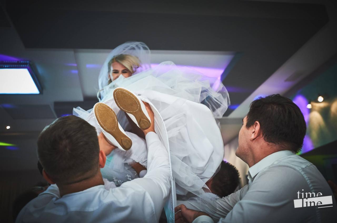 Uśmiechnięta panna młoda w białej sukni i welonie, podnoszona przez gości weselnych, widoczne podeszwy jej białych balerinek, oświetlenie sali weselnej w tle.