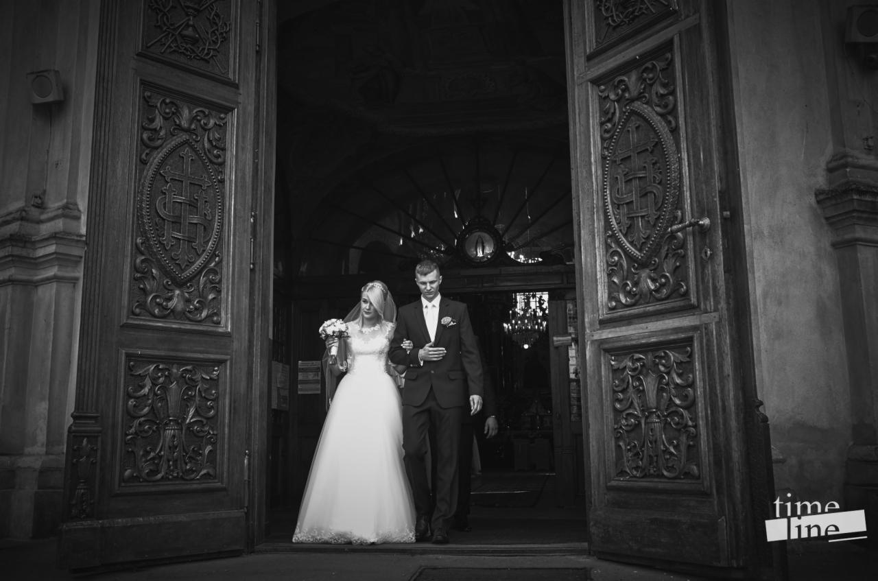 Czarno-białe zdjęcie nowożeńców wychodzących z kościoła przez ogromne, rzeźbione, drewniane drzwi, widoczny żyrandol wewnątrz, logo 'time line' w prawym dolnym rogu.