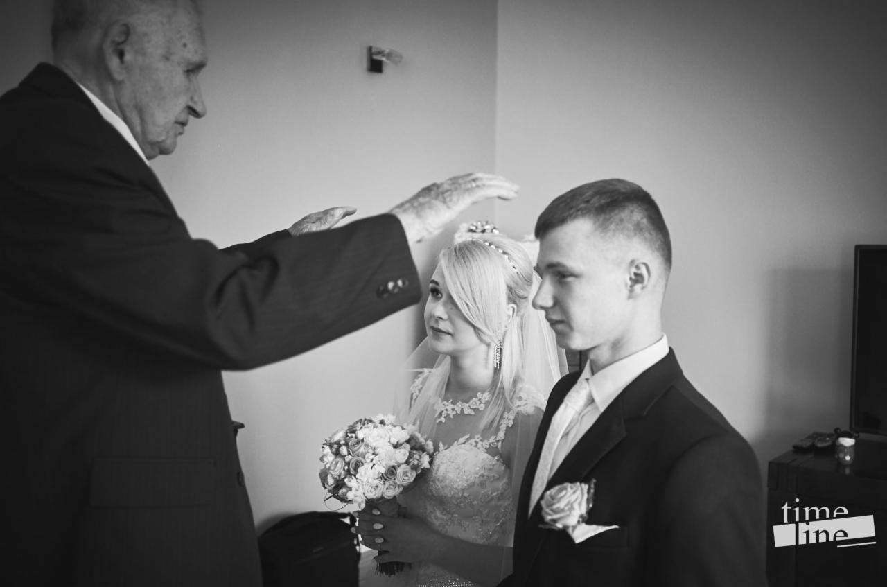 Czarno-białe zdjęcie panny młodej z bukietem i pana młodego w garniturze podczas błogosławieństwa przez starszego mężczyznę.