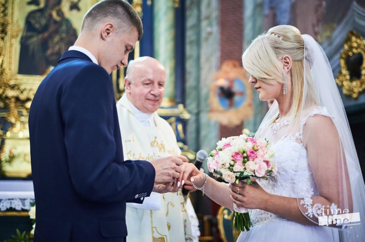 Para młoda wymienia obrączki podczas ceremonii ślubnej w kościele, w tle widoczny ksiądz