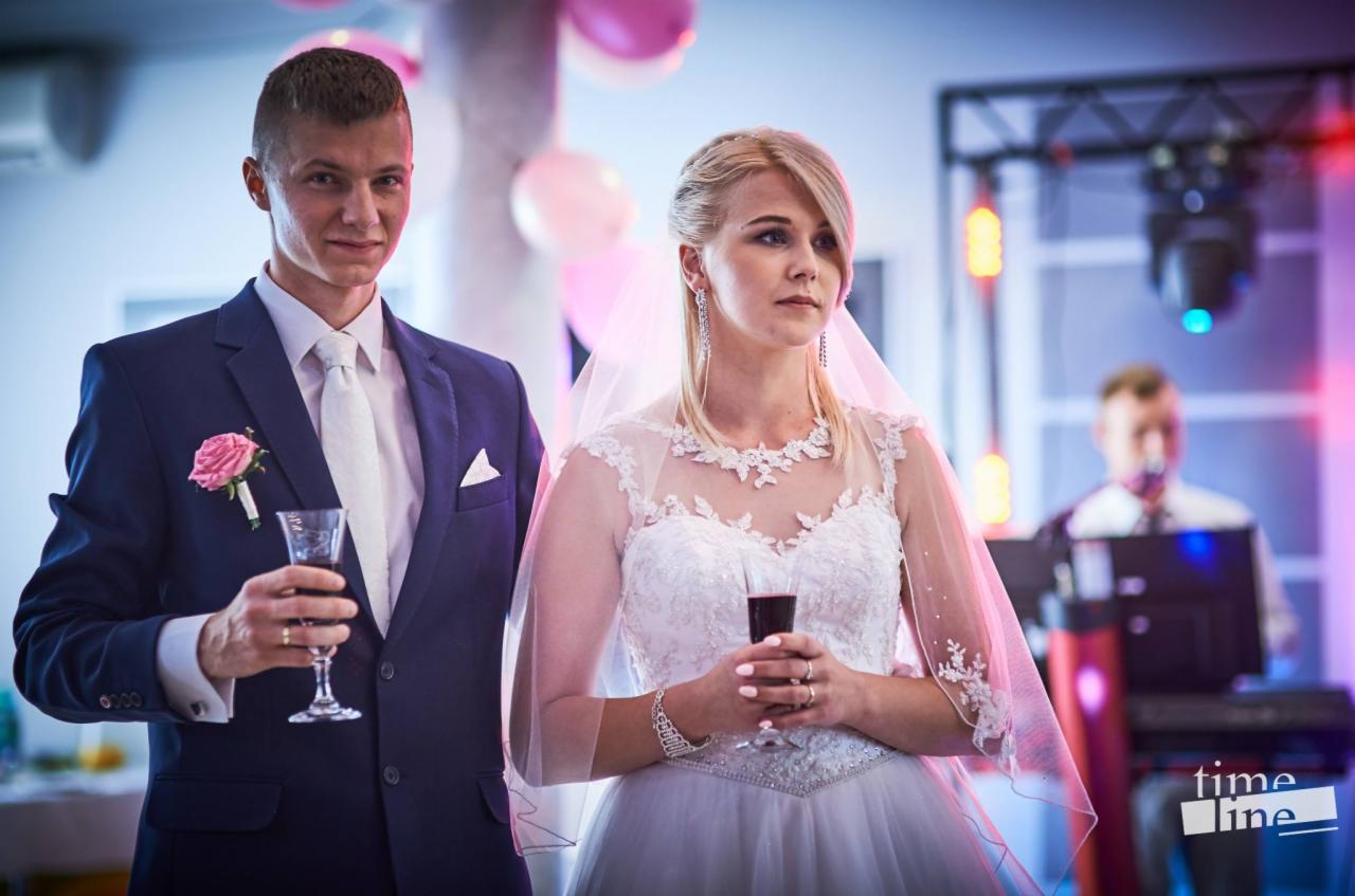 Para młoda w strojach ślubnych, on w granatowym garniturze z różową butonierką, ona w białej sukni z welonem, trzymają kieliszki z czerwonym winem na tle sceny z oświetleniem.