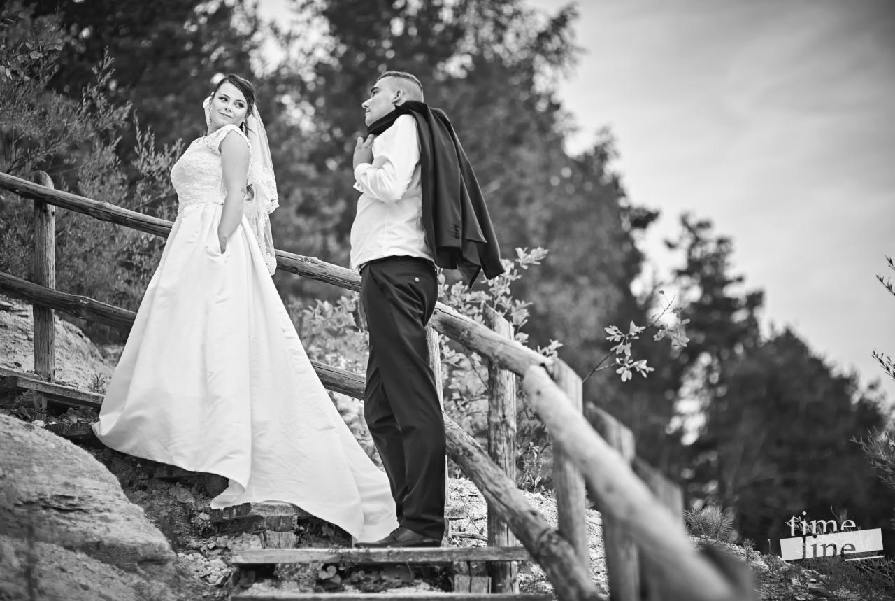 Czarno-białe zdjęcie pary młodej na drewnianych schodach w plenerze; panna młoda w sukni z koronkową górą i welonem, z rękami w kieszeniach, odwraca się; pan młody z marynarką przewieszoną przez...