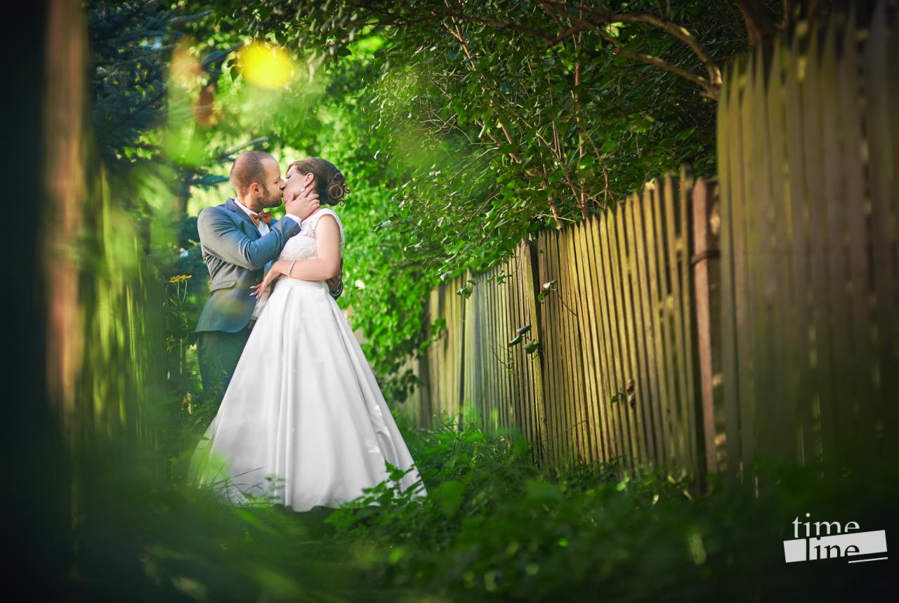 Para młoda w objęciach całuje się na tle drewnianego płotu i zieleni, w tle widoczne drzewa i logo fotografa w rogu kadru.