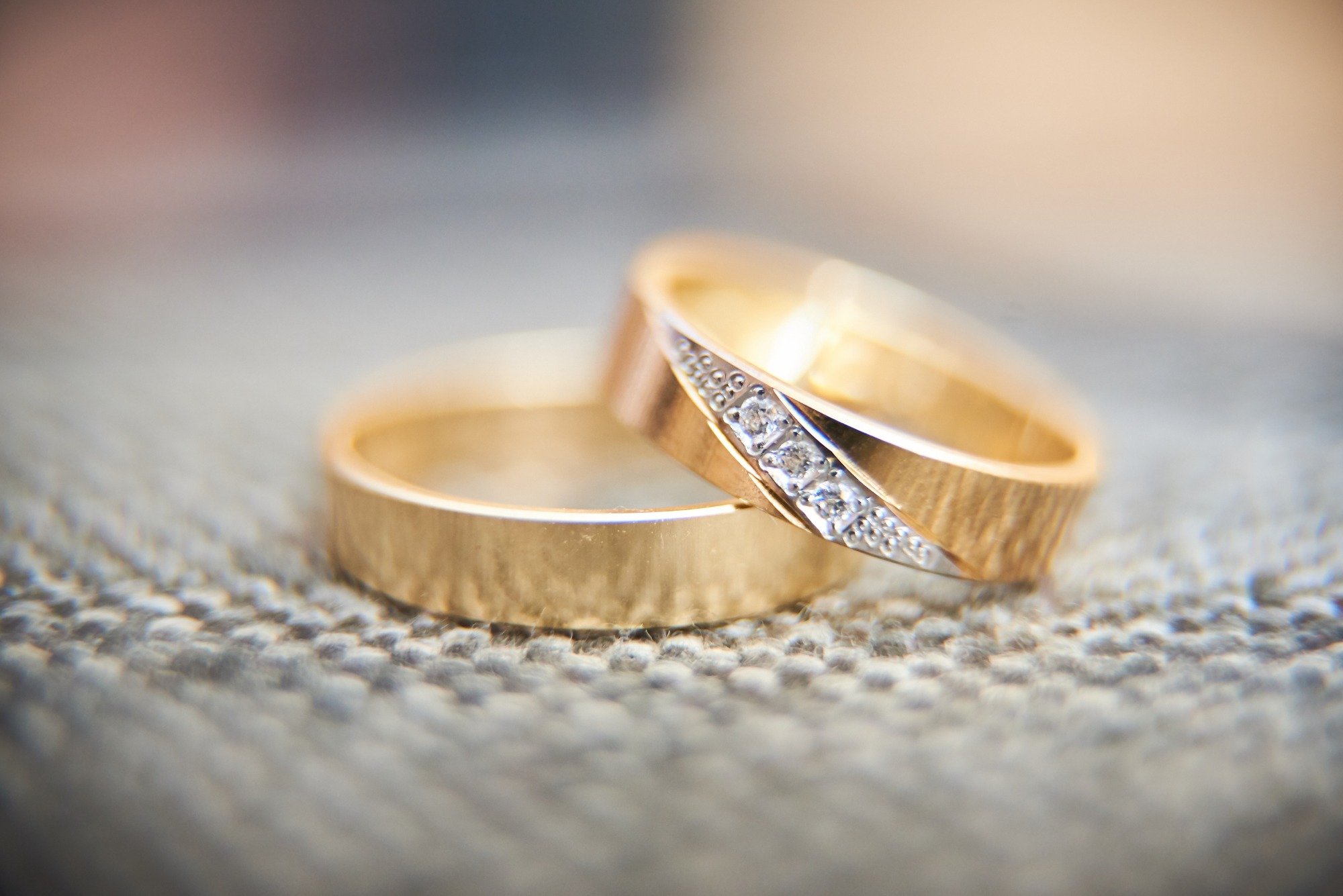 Dwa złote pierścionki ślubne leżące na teksturowanej, szarej powierzchni; jeden z pierścionków zdobiony rzędem drobnych diamentów.