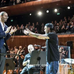 Dyrygent w niebieskiej marynarce i muzyk w czarnej koszulce oklaskują orkiestrę na scenie podczas koncertu, na widowni siedzą widzowie.