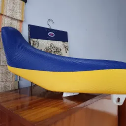 Odnowione, dwukolorowe (żółto-niebieskie) siedzisko motocyklowe z nową tapicerką, leżące na drewnianej komodzie, obok wzorniki tkanin tapicerskich.