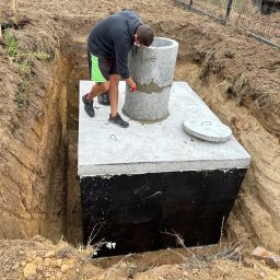 BULDMAX DAWID KOZŁOWSKI - Montaż betonowego szamba w wykopie w Czapurach. Widoczny pracownik uszczelniający połączenie kręgów betonowych. Ściany wykopu zabezpieczone przed osunięciem.