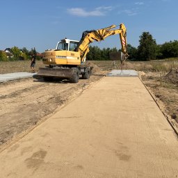 BULDMAX DAWID KOZŁOWSKI - Żółta koparka Liebherr na placu budowy, przygotowująca podłoże pod drogę, widoczny świeżo wyrównany piasek i płyty betonowe w tle.