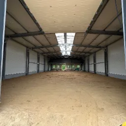 Wnętrze nowo wybudowanej hali z metalową konstrukcją, widoczne okna dachowe i przygotowane podłoże z ubitej ziemi, częściowo widoczny sprzęt budowlany w rogu.