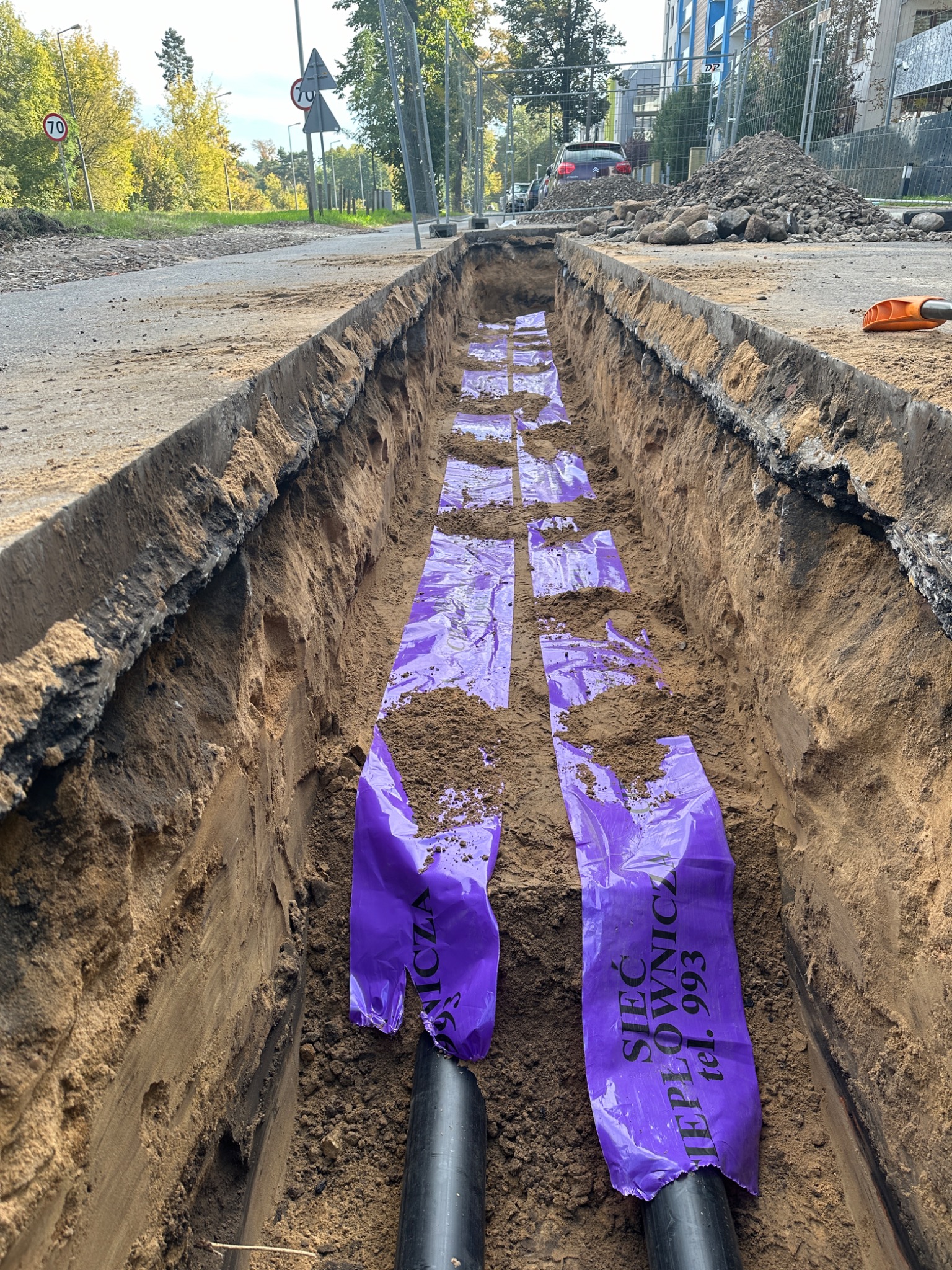 Głęboki wykop w ziemi wzdłuż drogi, w którym ułożone są czarne rury ciepłownicze zabezpieczone fioletową folią z napisem 'Sieć Ciepłownicza'. Widoczny znak drogowy ograniczenia prędkości do 70km/h...
