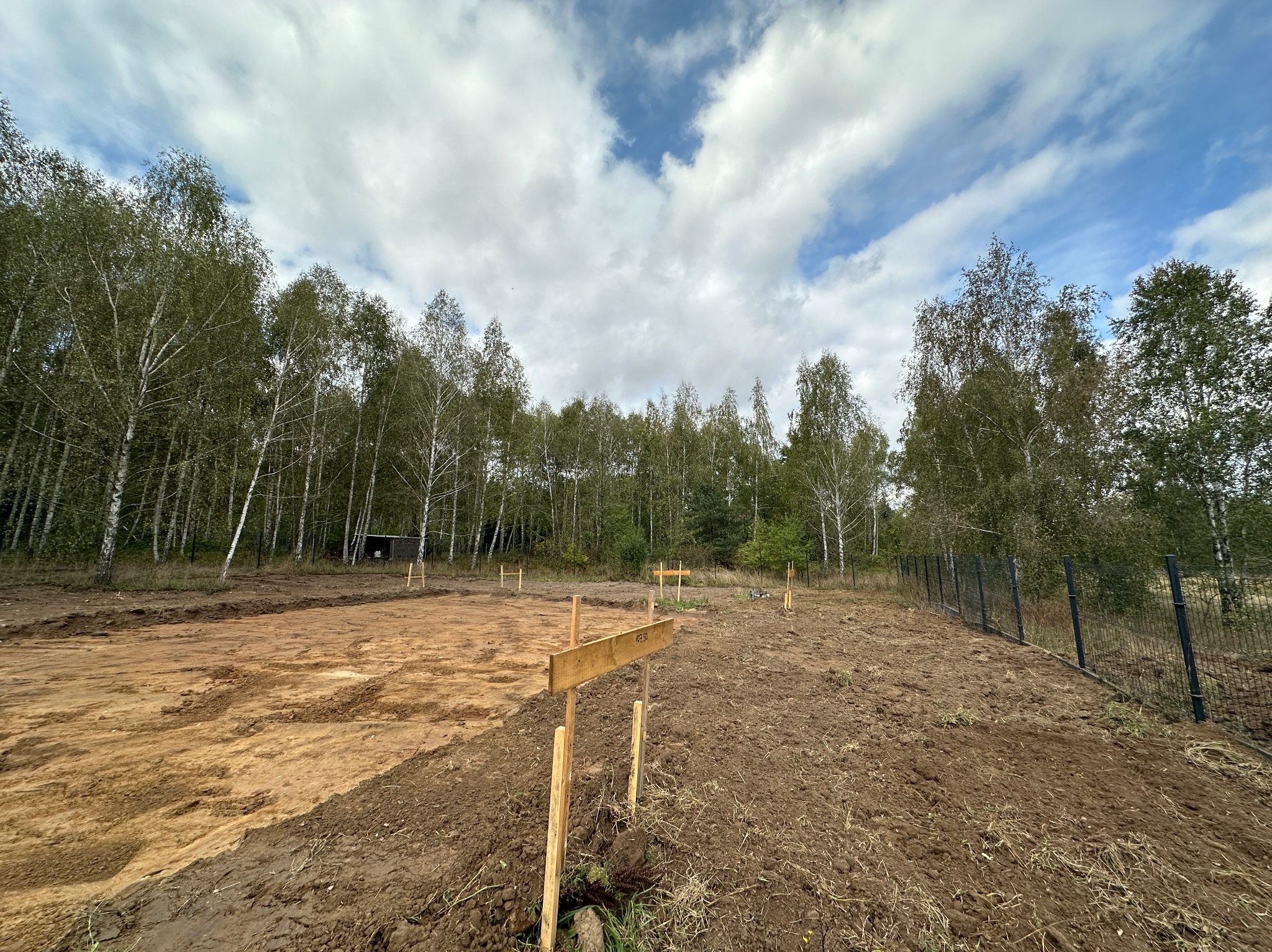 Przygotowany teren pod budowę z wytyczonymi punktami, widoczne drewniane słupki i deski, w tle las i ogrodzenie.