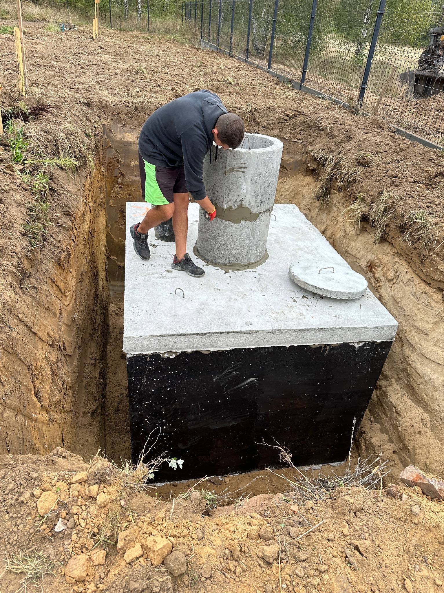 Montaż betonowego szamba w wykopie w Czapurach. Widoczny pracownik uszczelniający połączenie kręgów betonowych. Ściany wykopu zabezpieczone przed osunięciem.