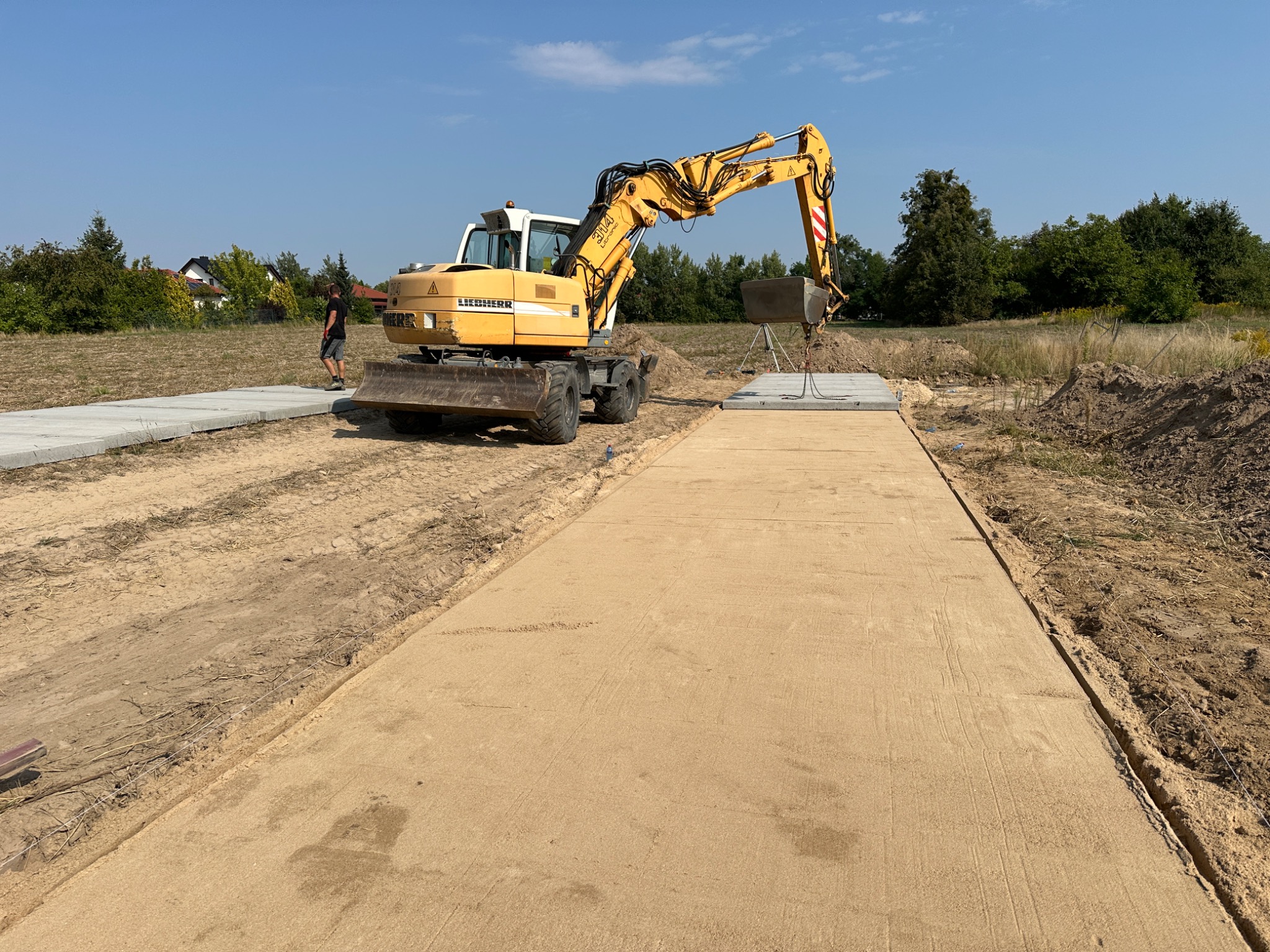 Żółta koparka Liebherr na placu budowy, przygotowująca podłoże pod drogę, widoczny świeżo wyrównany piasek i płyty betonowe w tle.