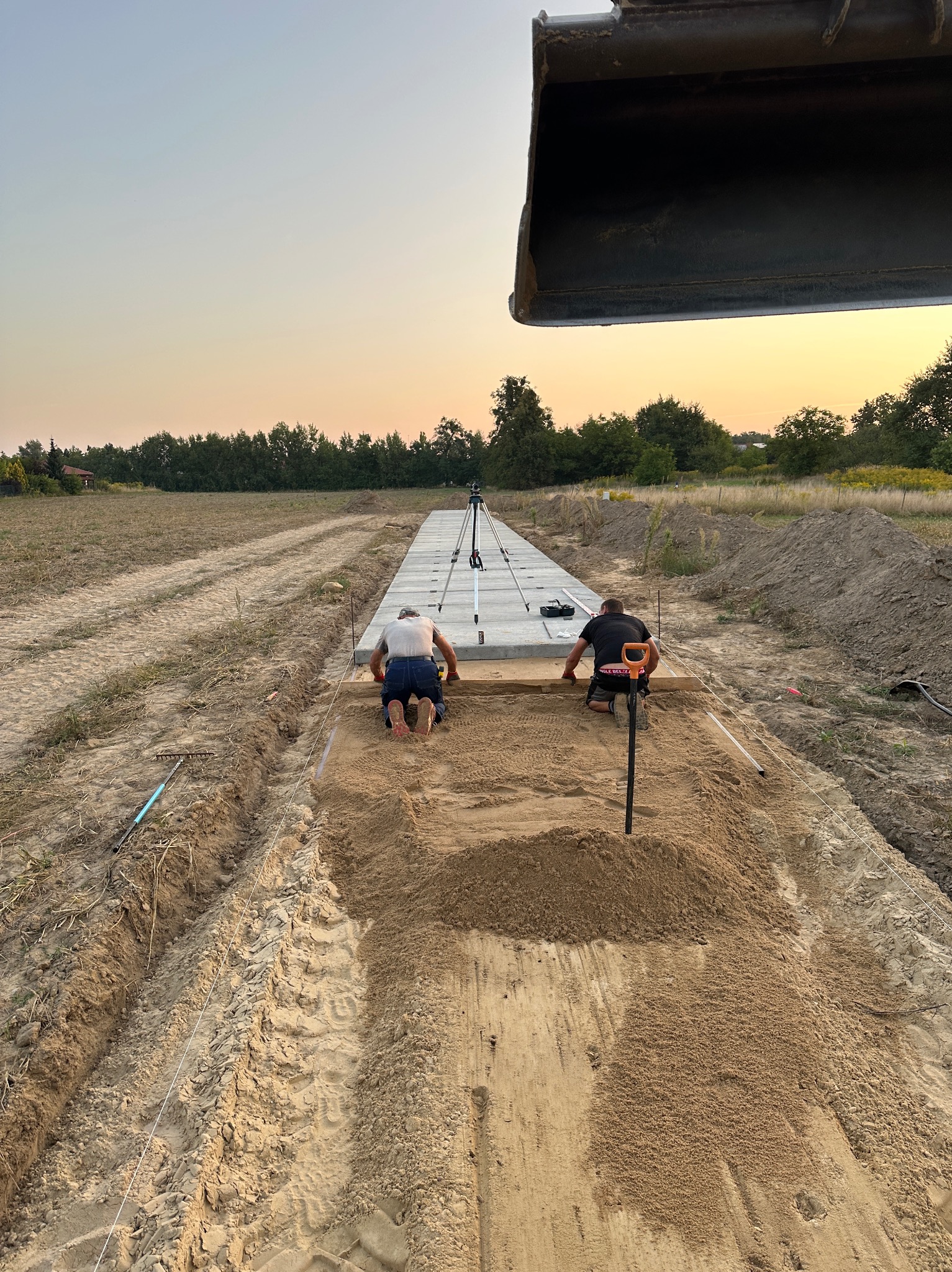 Dwóch mężczyzn klęczy na wyrównanym terenie, układając obrzeża pod przyszłą drogę lub chodnik; w tle widoczny krajobraz polny i ramię koparki.