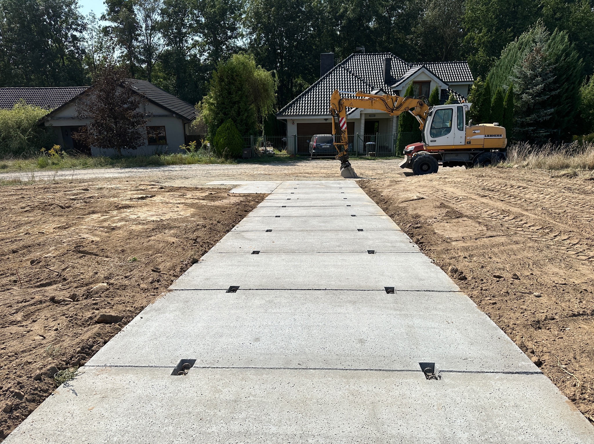 Ułożone betonowe płyty drogowe na przygotowanym terenie budowy z koparką Liebherr w tle, widok z perspektywy chodnika.