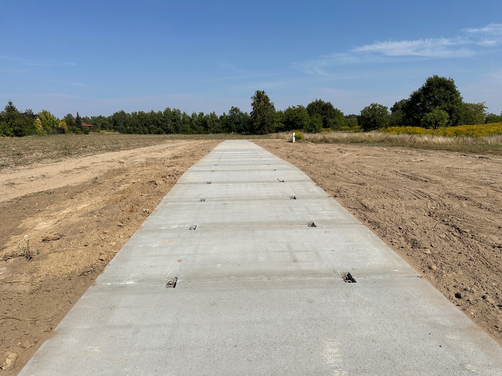 Utwardzona droga z betonowych płyt na przygotowanym terenie, widoczne metalowe uchwyty w płytach, tło z drzewami i błękitnym niebem.