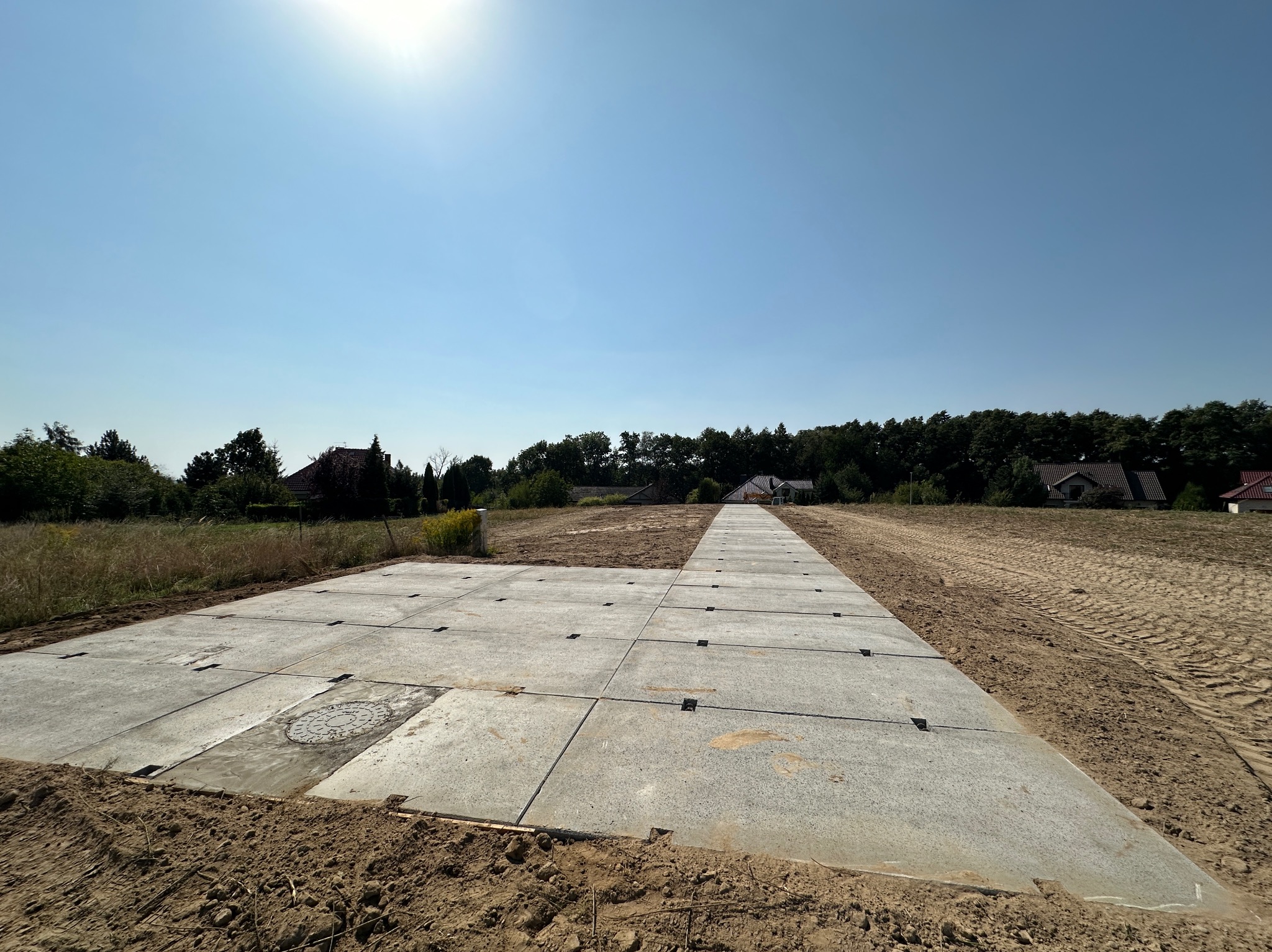 Przygotowany plac budowy z betonowymi płytami ułożonymi na piasku, widoczne domy w oddali pod błękitnym niebem.
