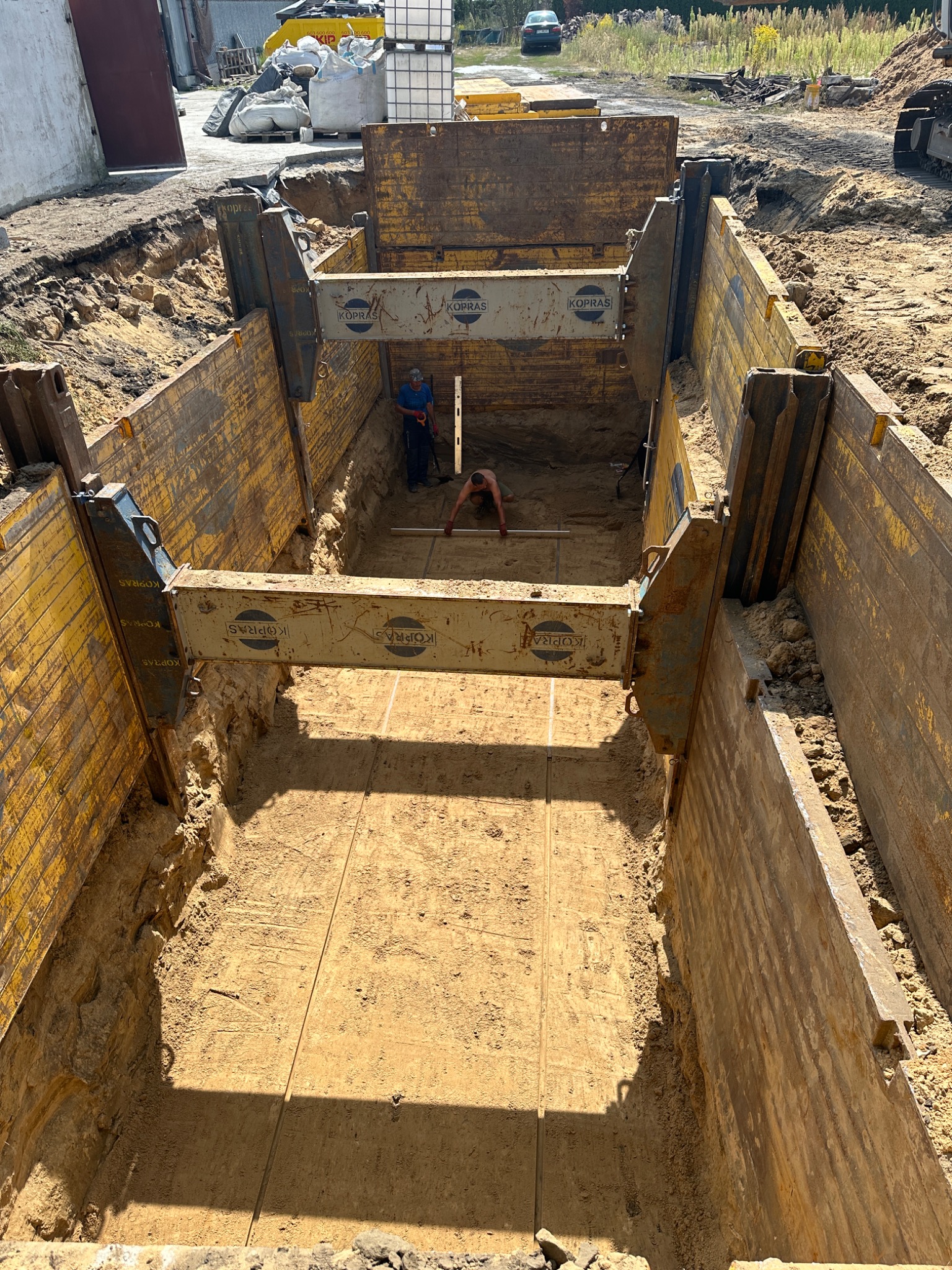 Głęboki wykop zabezpieczony metalowymi szalunkami, dwóch pracowników w środku, jeden z poziomicą, ziemia wokół wykopu, sprzęt budowlany w tle.