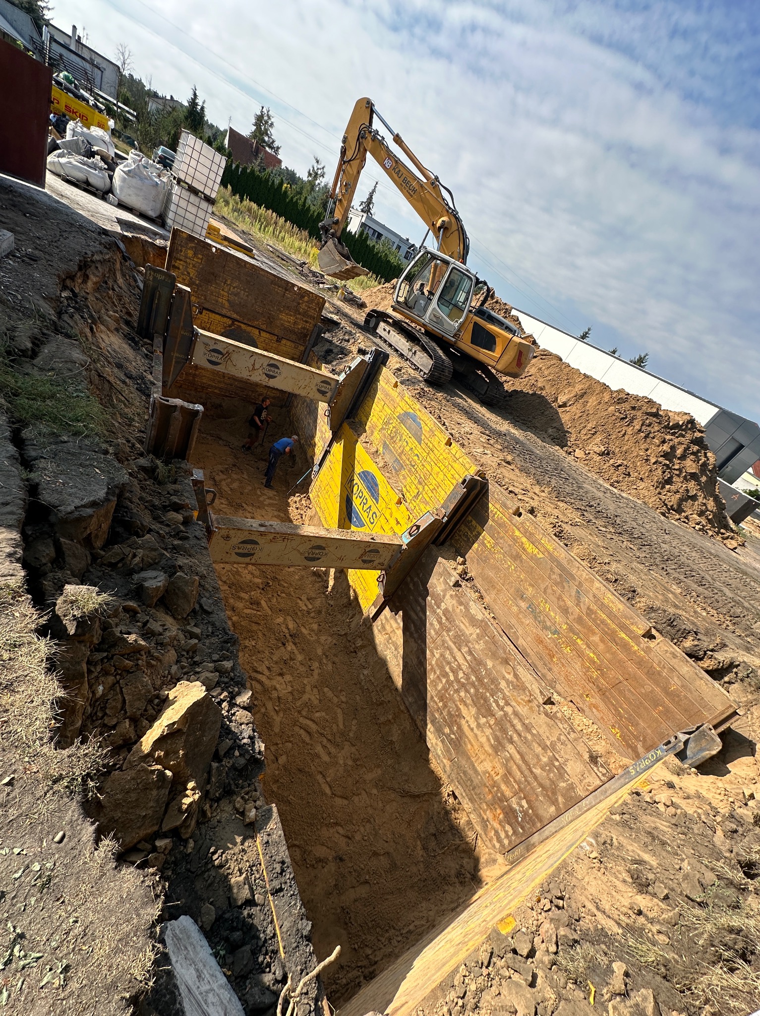 Żółte szalunki zabezpieczające wykop, wewnątrz dwóch pracowników, na górze koparka, w tle budynki i drzewa, ujęcie z góry pod kątem.