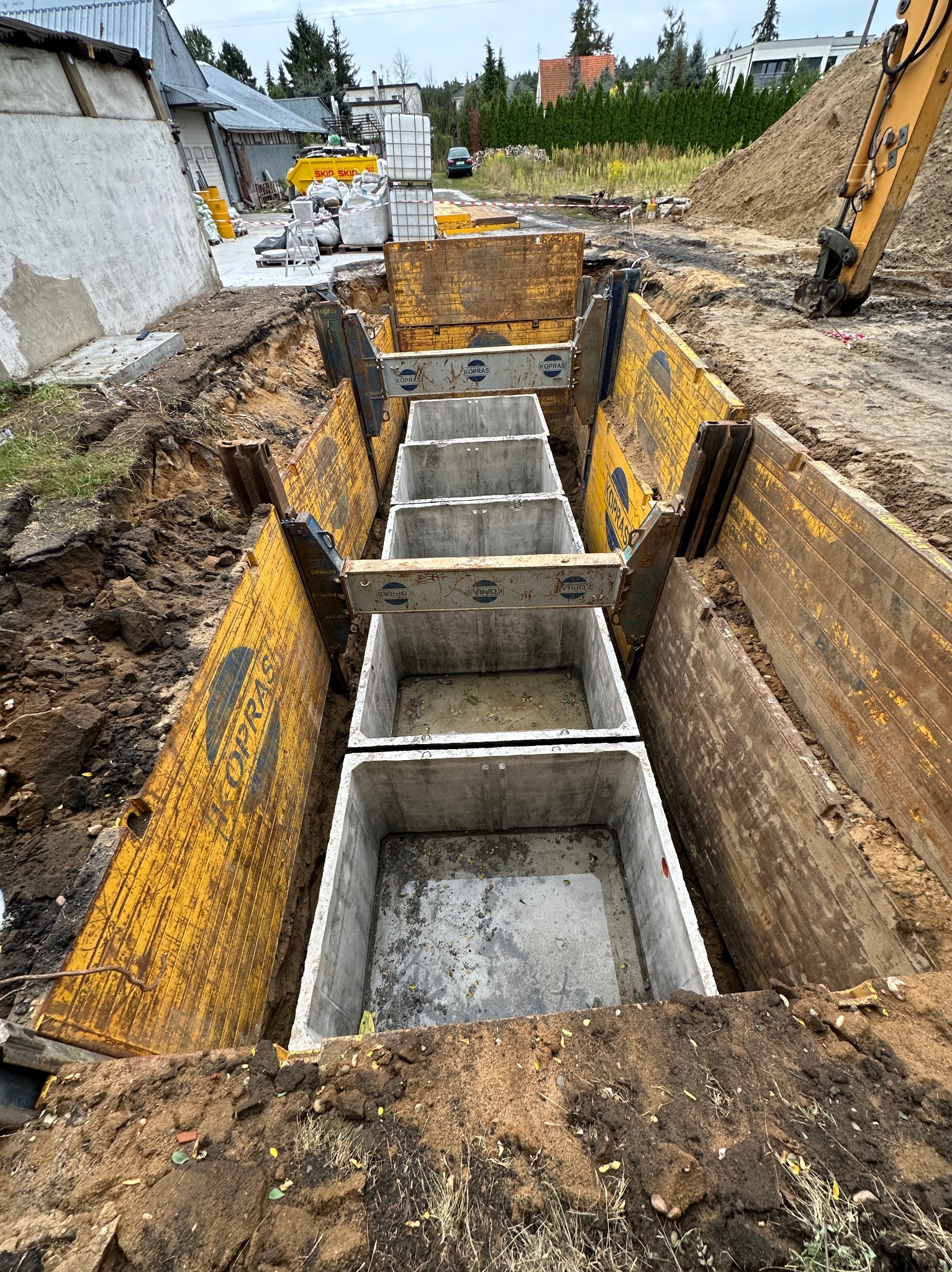 Głęboki wykop zabezpieczony żółtymi szalunkami KOPRAS, z ułożonymi prefabrykowanymi betonowymi elementami studni lub zbiornika; w tle widoczny budynek gospodarczy i koparka.