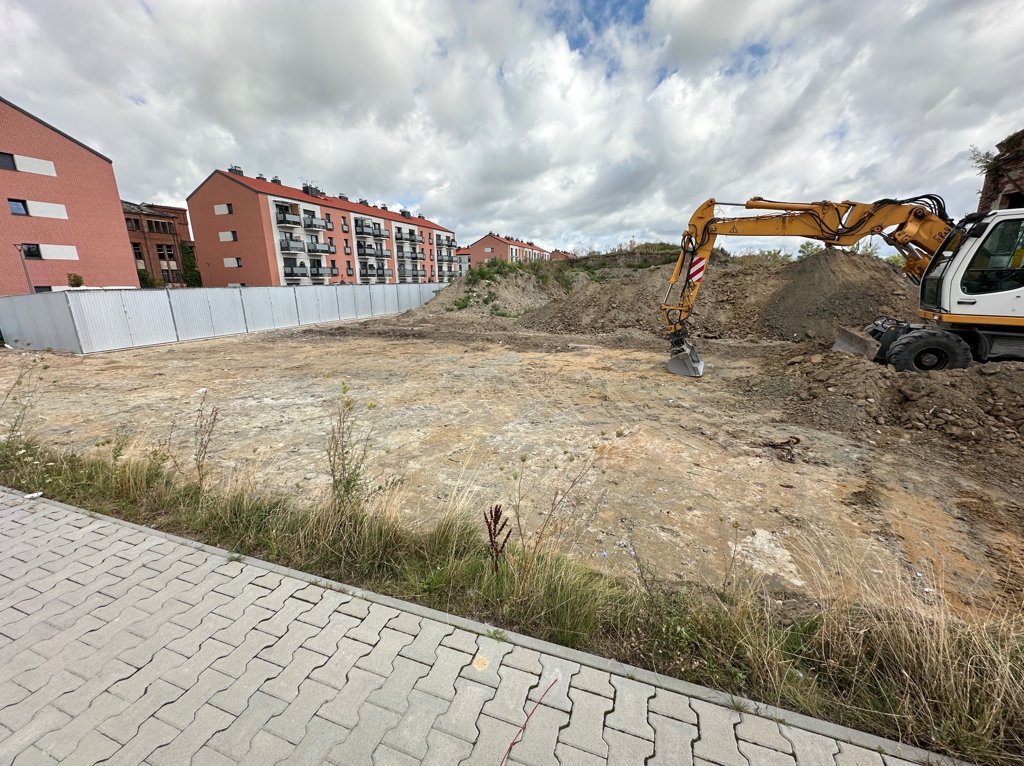 Żółto-biała koparka na placu budowy z pryzmą ziemi, w tle budynki mieszkalne i pochmurne niebo.