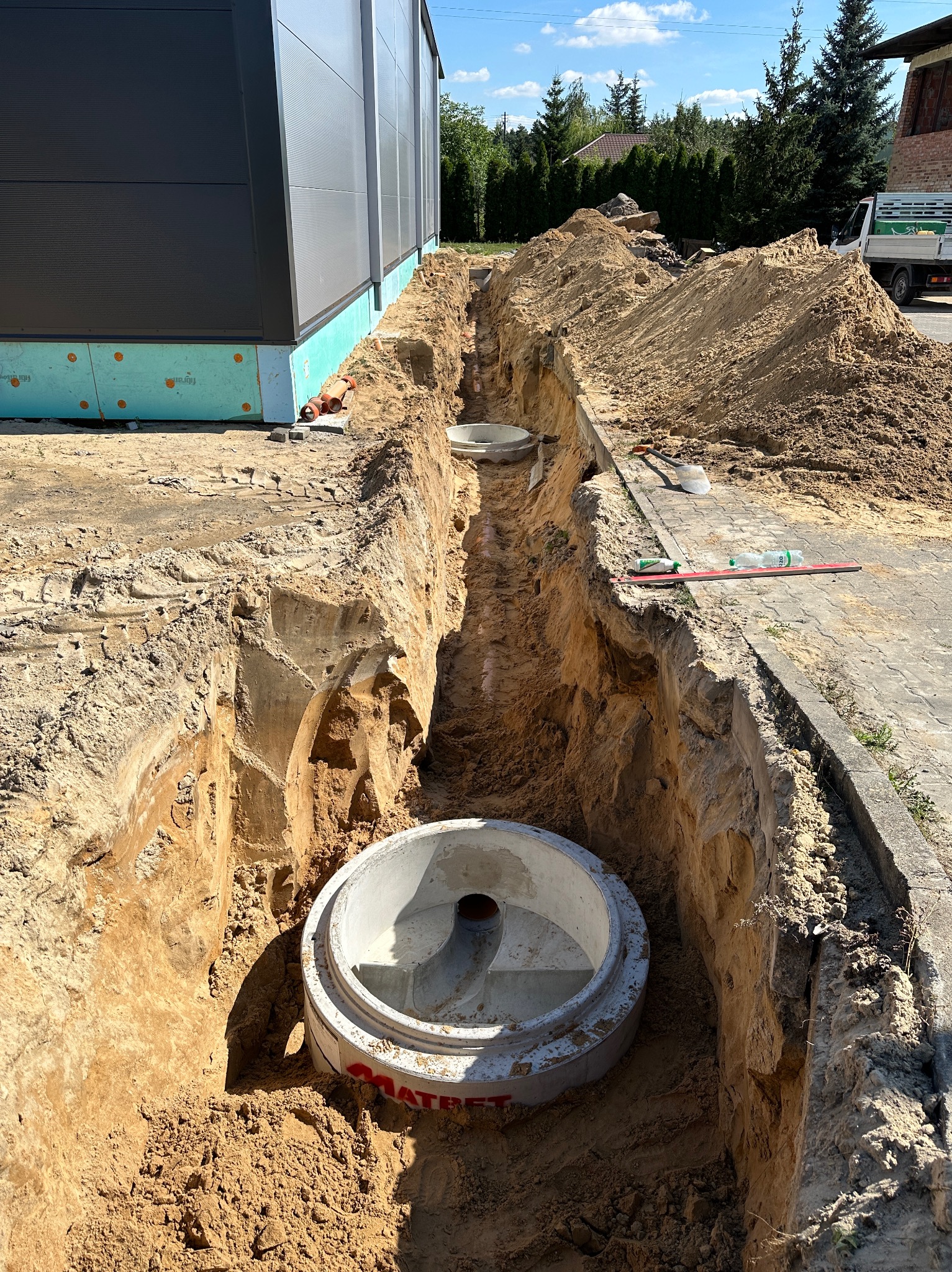 Głęboki wykop w ziemi z widocznymi betonowymi kręgami szamba, częściowo zamontowanymi, obok budynku przemysłowego z elementami izolacji.