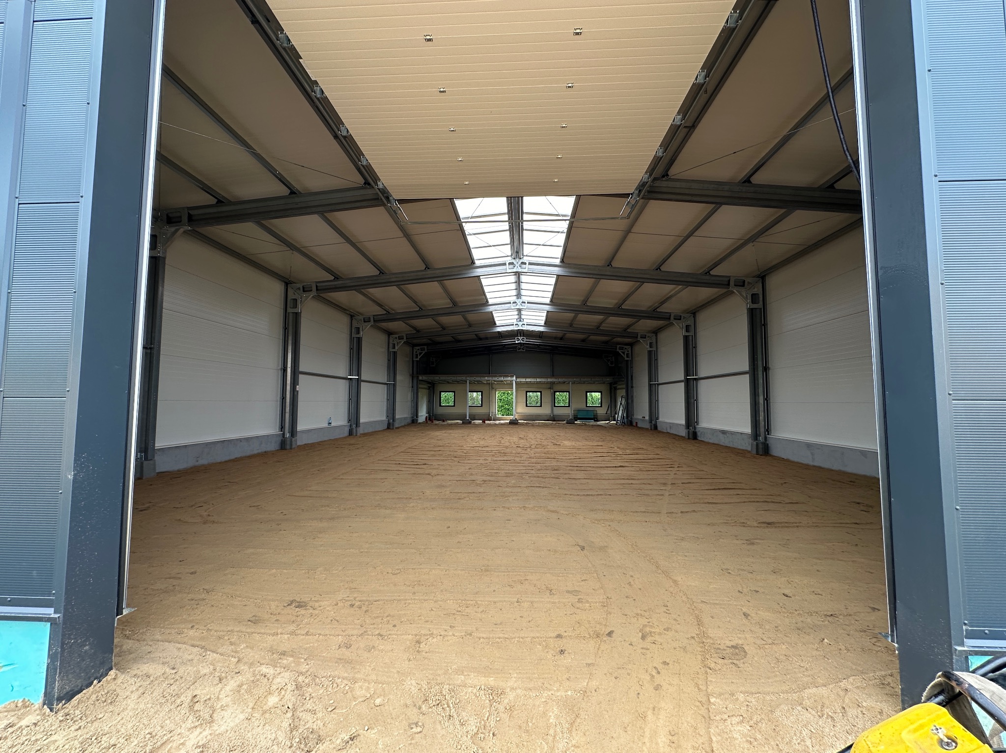 Wnętrze nowo wybudowanej hali z metalową konstrukcją, widoczne okna dachowe i przygotowane podłoże z ubitej ziemi, częściowo widoczny sprzęt budowlany w rogu.