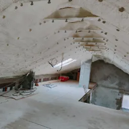 Poddasze po ociepleniu pianką PUR, widoczne elementy konstrukcyjne dachu i metalowe mocowania, fragment nieotynkowanej ściany z pustaków oraz prowizoryczna podłoga z płyt.