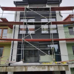 Wielopiętrowy budynek w trakcie prac elewacyjnych, widoczne rusztowania, fragmenty otynkowanej i pomalowanej fasady w kolorach bieli i czerni, wiadro z farbą na pierwszym planie.