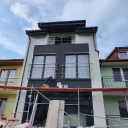 Pracownik na rusztowaniu maluje elewację nowoczesnego, trzypiętrowego budynku z czarno-białą fasadą, widok z dołu, na rusztowaniu puszki z farbą.