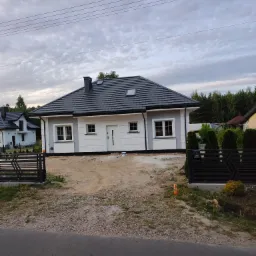 Nowo wybudowany parterowy dom jednorodzinny z szarą dachówką i białymi ścianami, widoczny z zewnątrz, z elementami wykończeniowymi w kolorze szarym, z czarnym metalowym ogrodzeniem w stylu...