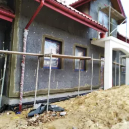 Dom w trakcie ocieplania, widoczne szare płyty izolacyjne na elewacji, okna zabezpieczone folią, rusztowania i czerwone rynny, teren wokół domu z ziemią i resztkami materiałów budowlanych.