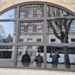 Effect Consulting-Krystian Stach - Arched window with brown frame reflecting a building and three people, showcasing the window's design and reflective properties.