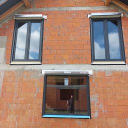 Effect Consulting-Krystian Stach - Ciemne okna z roletami podtynkowymi w surowej, ceglanej ścianie budynku w trakcie budowy. W dolnym oknie odbija się sylwetka osoby.