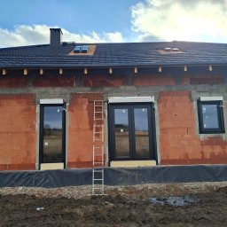 Effect Consulting-Krystian Stach - Surowy mur budynku z czarnymi oknami i roletami podtynkowymi w trakcie montażu. Na ścianie oparta drabina. Widoczne fragmenty nieba z chmurami.