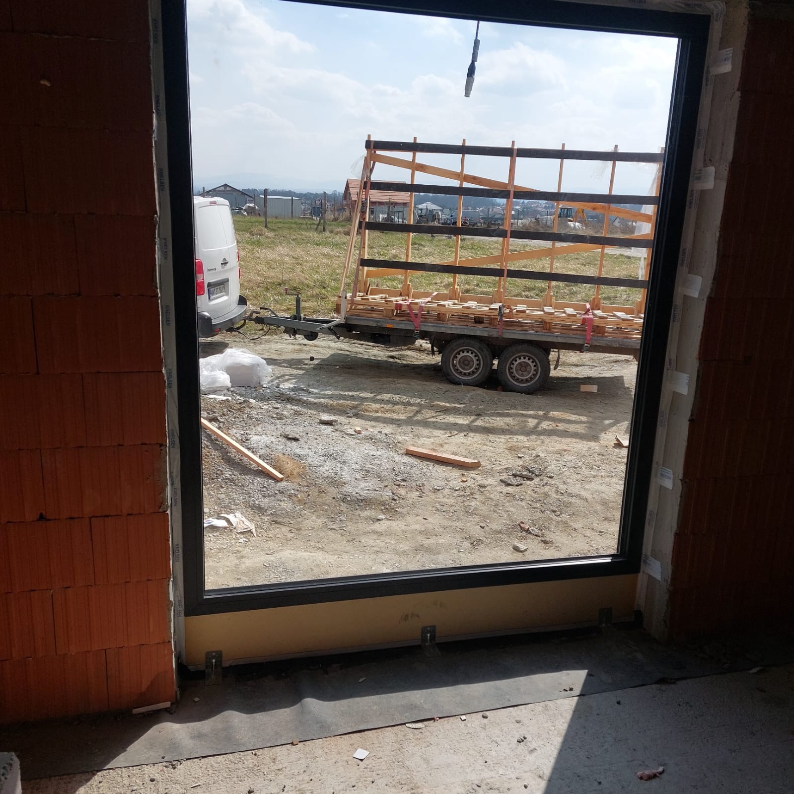 Czarne okno PCV w surowym murze, widok na przyczepę z oknami i biały samochód dostawczy na zewnątrz. Inwestycja w trakcie realizacji, profil Salamander.