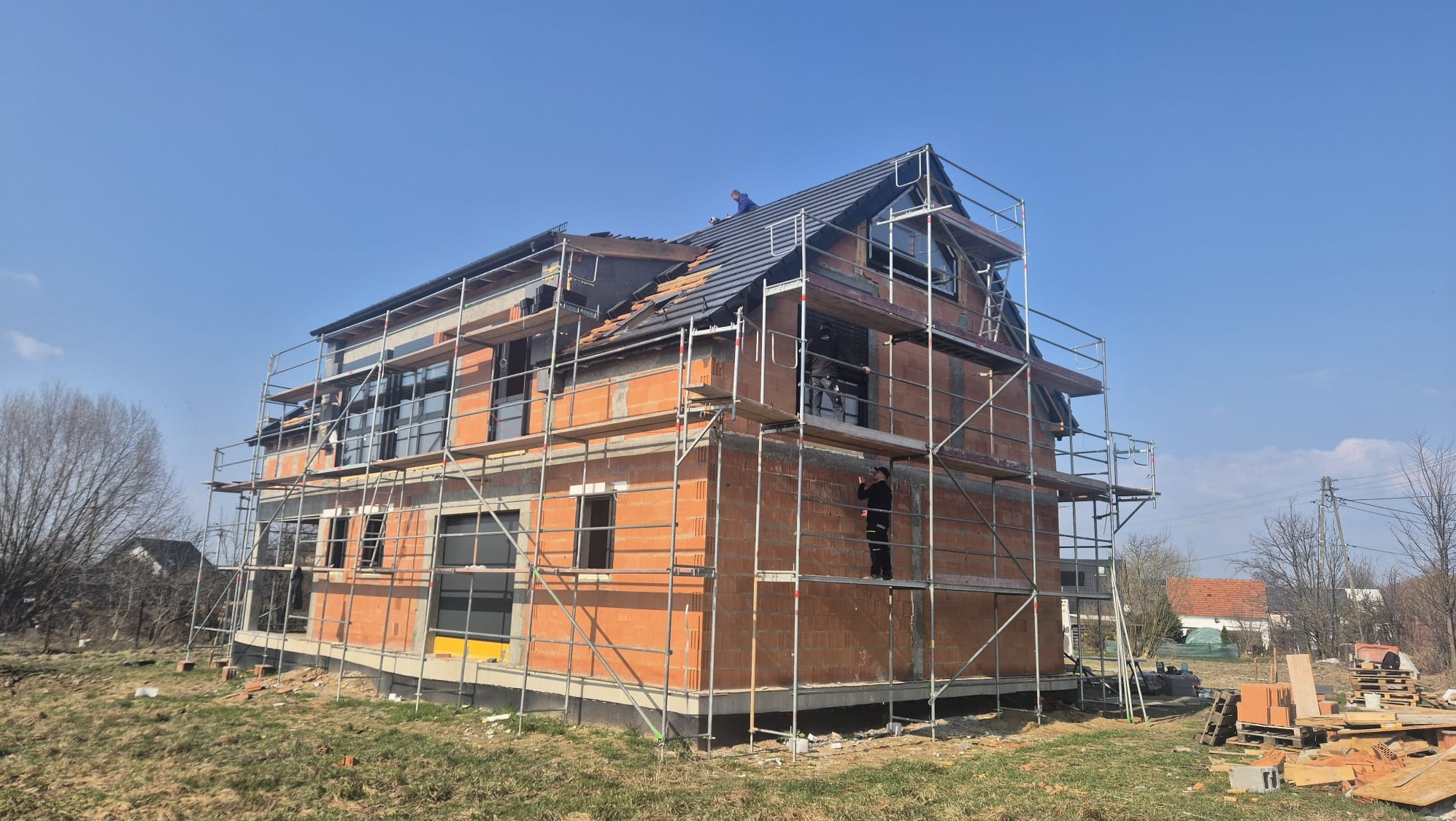 Budowa domu jednorodzinnego w trakcie montażu okien i układania dachu. Widoczne rusztowania, cegły, okna wstawione w otwory, pracownicy na dachu i rusztowaniach.