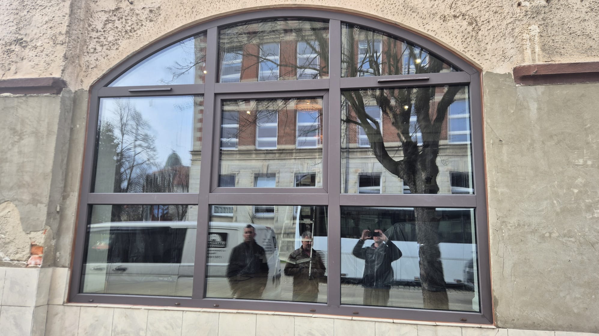 Arched window with brown frame reflecting a building and three people, showcasing the window's design and reflective properties.