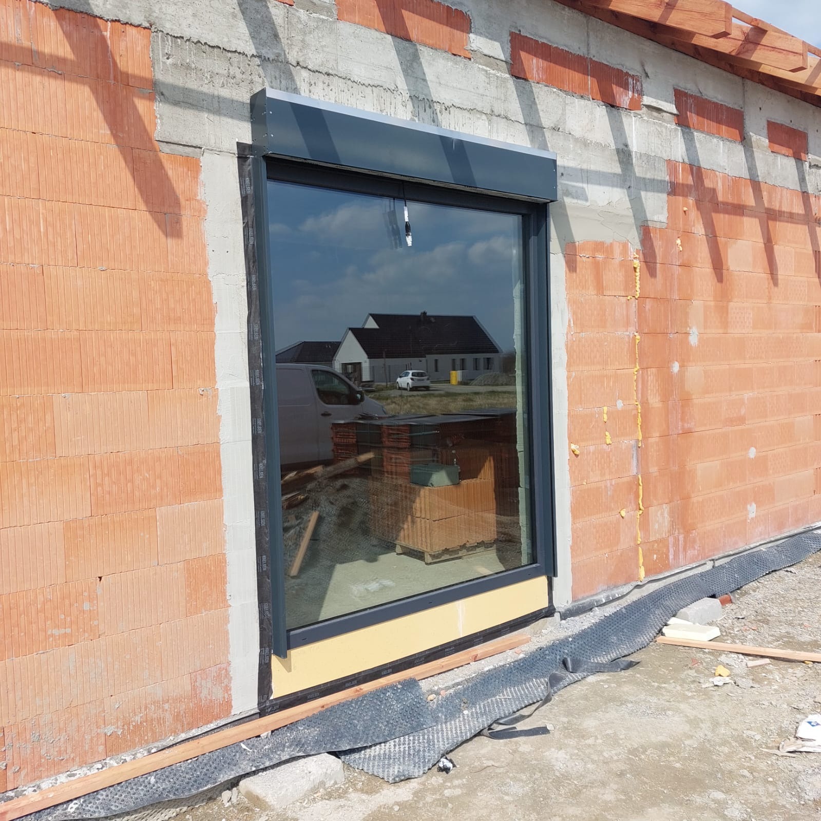 Szare okno z zamontowaną roletą zewnętrzną w surowym murze z cegły. Widoczne detale montażu i odbicie otoczenia w szybie.