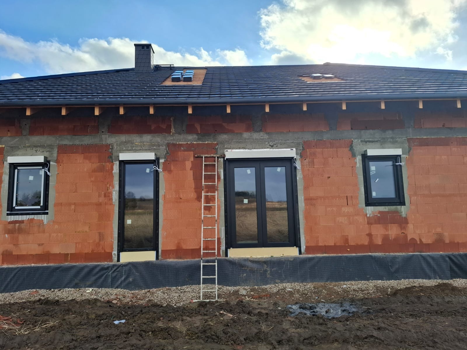 Surowy mur budynku z czarnymi oknami i roletami podtynkowymi w trakcie montażu. Na ścianie oparta drabina. Widoczne fragmenty nieba z chmurami.