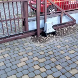 Brązowa brama przesuwna z automatyką, widoczny napęd zamontowany na fundamencie, kostka brukowa na podjeździe oraz fragment czerwonego samochodu w tle.