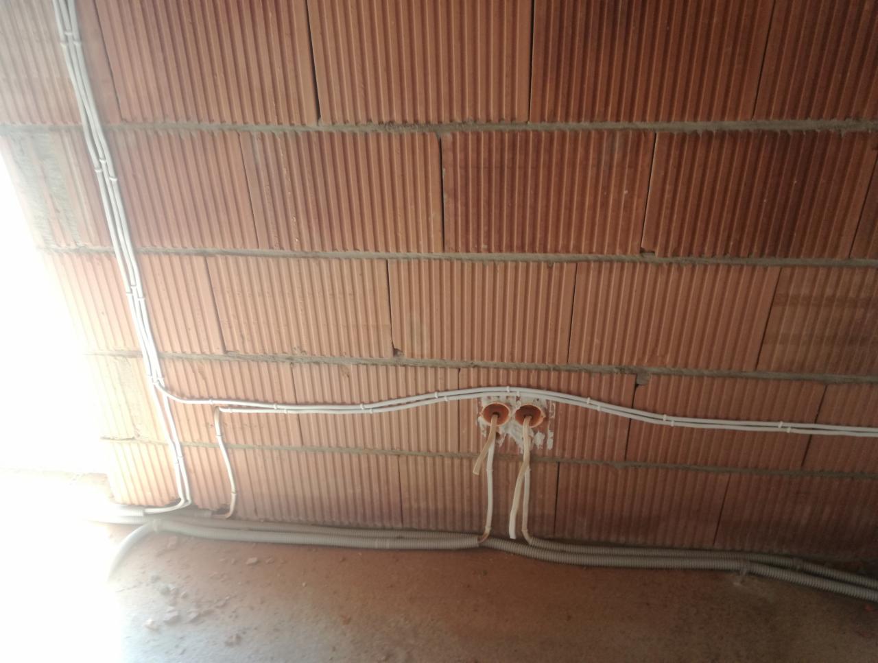 Instalacja przewodów elektrycznych w białych rurkach na ceglanym suficie podwieszanym, widoczne puszki instalacyjne i peszel przy podłodze.