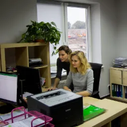 Dwie kobiety w biurze, jedna stoi i patrzy na ekran komputera, druga siedzi, na biurku drukarka Brother i dokumenty, za oknem widok na plac budowy z maszynami.