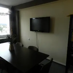 Sala konferencyjna lub klasa z dużym stołem, krzesłami, telewizorem na ścianie i regałem z książkami, w tym podręcznikiem do TOEFL, z widokiem na miasto za oknem.